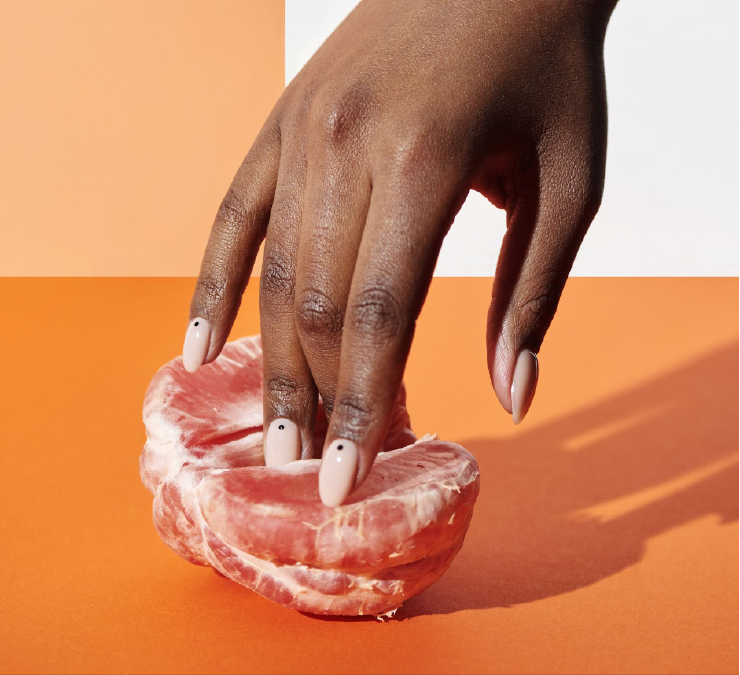 A photo of half of a peeled grapefruit. A dark skinned hand with pale pink painted nails has put their fingers in between the segment of the grapefruit giving a ‘suggestive’ quality to the image.