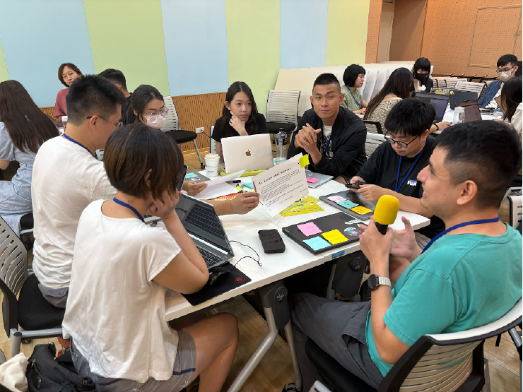 A group of 7 Taiwanese designers are gathered around a table with laptops, papers and sticky notes on the table. One person holds a microphone and speaks about the project that the group decided to work on making contributions to. In the background, two other tables of designers are visible