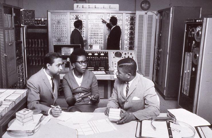 A group of Census Bureau researches talking and making notes behind a full room of 50’s era computer machinery.