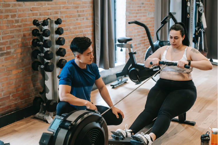A masc presenting exercise coach and a femme presenting person using a rowing machine in exercise clothing. The coach is watching how they position the rowing bar and supporting them. Image chosen for this blog subject due to the metaphor of how coaching is like supporting people in their own training and own ability to strengthen and overcome their project challenges.