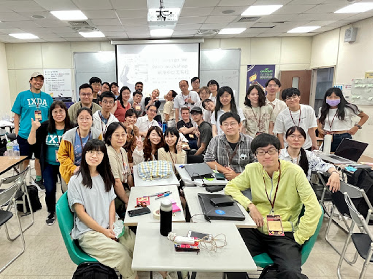 A photo of “The Design We Open” workshop in Taiwan at COSCUP 2023. There are 20 plus people gathered around a table with laptops, sticky notes and writing on the tables from the workshop. Everyone is looking towards the camera and some people are smiling.